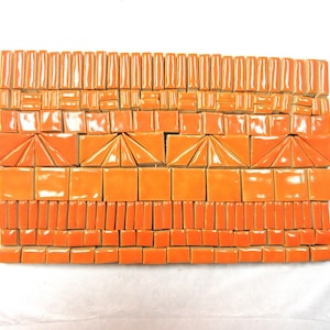 May include: A collection of bright orange ceramic mosaic tiles arranged in a rectangular grid. The set includes various shapes such as small rectangles, squares, and triangles with a glossy finish, showcasing a uniform vibrant orange color.