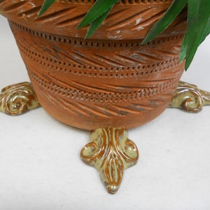 May include: A terracotta-colored ceramic planter with carved geometric patterns and decorative feet. The planter has a rounded shape and is supported by three ornate, light-greenish-brown feet. Green plant leaves are visible at the top.