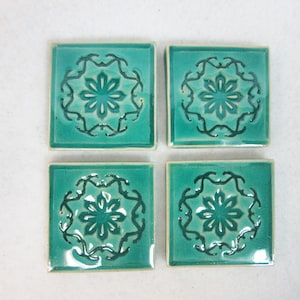 May include: Four square ceramic tiles with a teal glaze. Each tile features a dark teal floral design in the center, surrounded by a decorative border. The tiles are arranged in a square formation, showcasing their decorative potential.