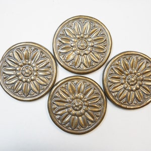 May include: Four gold-colored metal coasters with a raised floral design. Each coaster features a circular design with a central flower motif.