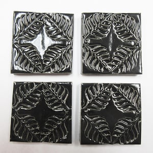 May include: Four square, black tiles with a raised, silver leaf design. Each tile features a central diamond shape with a cross-like pattern. The tiles are arranged in a square formation against a white background.