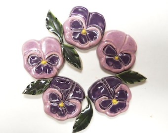 May include: A collection of five ceramic pansy flower ornaments arranged in a circular pattern. Each flower features glossy light purple and dark purple petals with a small yellow center, accompanied by individual dark green ceramic leaves.