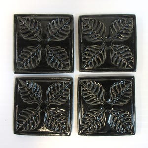 May include: Four square, black ceramic coasters. Each coaster features a raised leaf design in a symmetrical pattern. The coasters have a glossy finish and are arranged in a square formation, perfect for protecting surfaces from drink rings.