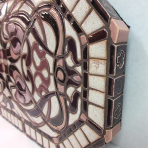 May include: Close-up of a ceramic mosaic wall art piece. The artwork features an intricate design in shades of mauve, cream, and burgundy. The mosaic tiles are arranged in a geometric pattern, creating a visually appealing texture. The piece has a unique octagonal shape with a defined border. The craftsmanship is evident in the precise placement of each tile, resulting in a high-quality, decorative piece suitable for interior design.