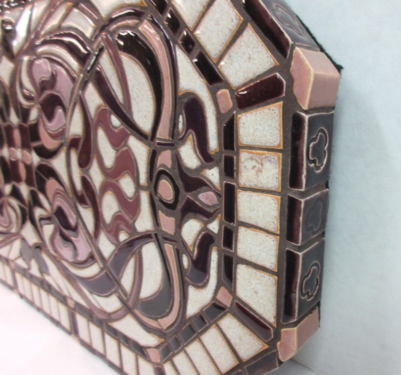 May include: Close-up of a ceramic mosaic wall art piece. The artwork features an intricate design in shades of mauve, cream, and burgundy. The mosaic tiles are arranged in a geometric pattern, creating a visually appealing texture. The piece has a unique octagonal shape with a defined border. The craftsmanship is evident in the precise placement of each tile, resulting in a high-quality, decorative piece suitable for interior design.