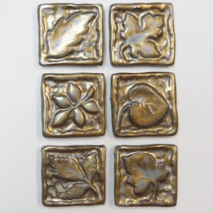 May include: Six square ceramic tiles with a raised design of different leaf shapes. The tiles are a metallic gold color with a silver glaze.