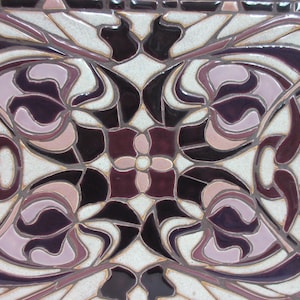 May include: Close-up view of a ceramic tile showcasing an intricate design. The tile features a symmetrical pattern in shades of purple, pink, and off-white. The design incorporates stylized floral motifs and flowing lines, creating a sense of elegance and sophistication. The tile's surface appears glossy, reflecting light. The color palette is predominantly dark purple and light pink, with the off-white acting as a background. The design is reminiscent of Art Nouveau or Art Deco styles.