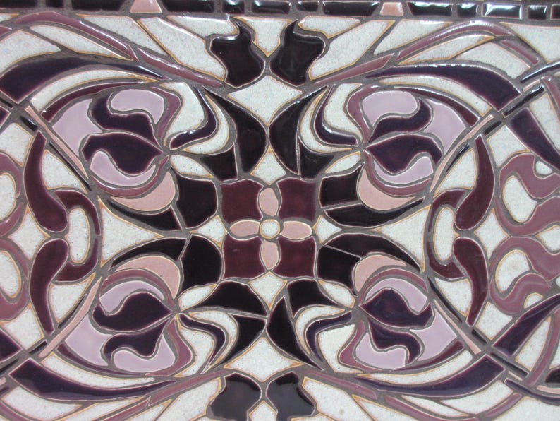 May include: Close-up view of a ceramic tile showcasing an intricate design. The tile features a symmetrical pattern in shades of purple, pink, and off-white. The design incorporates stylized floral motifs and flowing lines, creating a sense of elegance and sophistication. The tile's surface appears glossy, reflecting light. The color palette is predominantly dark purple and light pink, with the off-white acting as a background. The design is reminiscent of Art Nouveau or Art Deco styles.