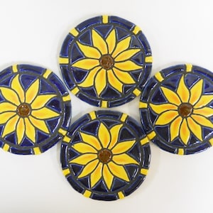 May include: Set of four ceramic coasters featuring a sunflower design. Each coaster has a blue background with a yellow sunflower in the center. The sunflowers have a brown center and yellow petals.