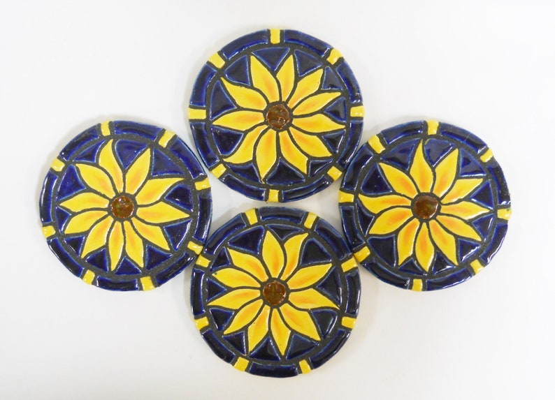 May include: Set of four ceramic coasters featuring a sunflower design. Each coaster has a blue background with a yellow sunflower in the center. The sunflowers have a brown center and yellow petals.