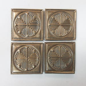 May include: Four square, bronze-colored tiles arranged in a square. Each tile features a circular design with a cross-like pattern at the center, surrounded by geometric lines. The tiles have a slightly raised, textured surface and a metallic sheen.