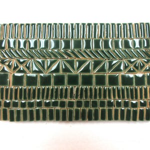 May include: A collection of dark forest green ceramic tiles arranged in a rectangular grid. The tiles feature various shapes including small squares, thin rectangles, and triangles, all displaying a glossy finish with visible light reflections.