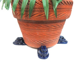 May include: A large, terracotta-colored ceramic plant pot with intricate carved patterns. The pot rests on three small, dark blue ceramic feet. Green plant leaves are visible at the top of the pot.