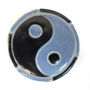 May include: A circular ceramic tile featuring a glossy yin and yang symbol. The design consists of two interlocking teardrop shapes in solid black and muted blue glaze, each containing a small circular dot of the contrasting color in the center.