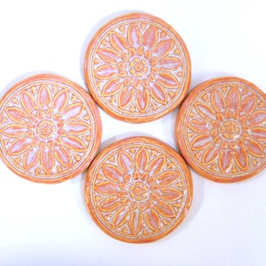 May include: Four ceramic coasters with a white and orange floral design. The coasters are round and have a raised design.
