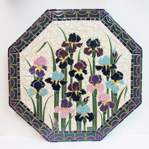 May include: Octagonal ceramic tile with a mosaic design of purple, blue, and pink iris flowers on a white background. The tile has a green and purple border.