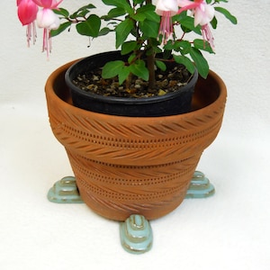 May include: A terracotta flower pot with a decorative design sits on a set of three teal blue ceramic feet. The pot is filled with soil and a small plant with pink flowers.