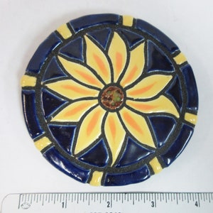 May include: A round ceramic tile with a blue background and a yellow flower design. The flower has orange accents and a brown center.