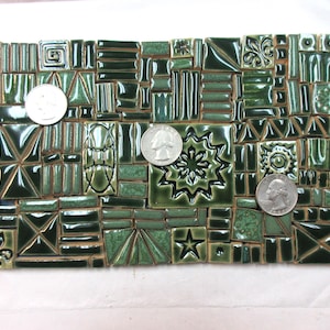 May include: A collection of dark green and sage green ceramic mosaic tiles in various geometric shapes, including squares, rectangles, and triangles, featuring embossed patterns like stars and fleur-de-lis, with three US quarters for scale.