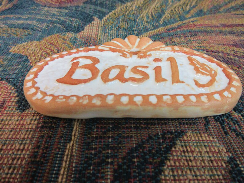 May include: A white ceramic kitchen herb label with a brown border and the word "Basil" written in brown lettering.