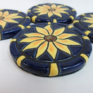 May include: Four ceramic coasters with a blue background and a yellow flower design. The flowers have a yellow center with orange accents.