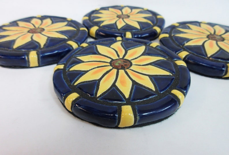 May include: Four ceramic coasters with a blue background and a yellow flower design. The flowers have a yellow center with orange accents.