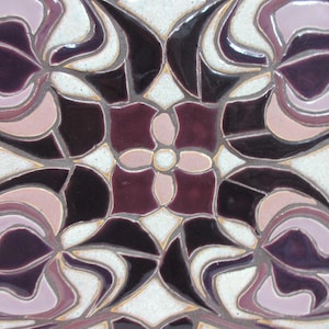 May include: Close-up view of a ceramic tile showcasing an intricate design. The tile features a symmetrical pattern of stylized floral motifs in shades of deep purple, mauve, and off-white. The design incorporates curved lines and geometric shapes, creating a sense of depth and movement. The colors are rich and saturated, with a glossy finish that enhances the visual appeal. The tile's surface appears smooth and polished, suggesting a high-quality craftsmanship.