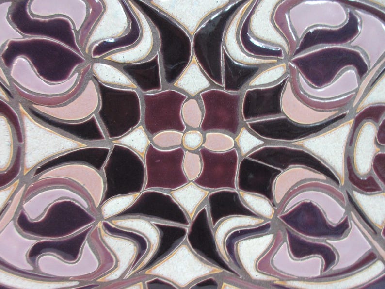 May include: Close-up view of a ceramic tile showcasing an intricate design. The tile features a symmetrical pattern of stylized floral motifs in shades of deep purple, mauve, and off-white. The design incorporates curved lines and geometric shapes, creating a sense of depth and movement. The colors are rich and saturated, with a glossy finish that enhances the visual appeal. The tile's surface appears smooth and polished, suggesting a high-quality craftsmanship.