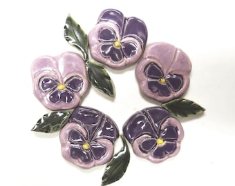 May include: A collection of five ceramic pansy flower ornaments arranged in a circular pattern. Each flower features glossy light purple and dark purple petals with a small yellow center, accented by several dark green ceramic leaves.