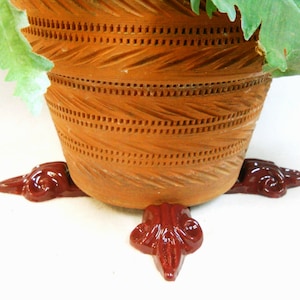 May include: A terracotta flower pot with a decorative pattern sits on three red, decorative feet. The pot is filled with green leafy plants.