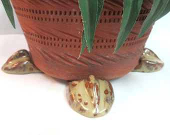May include: A terracotta planter with a woven design and four decorative feet. The planter has a brown glaze with a speckled pattern.