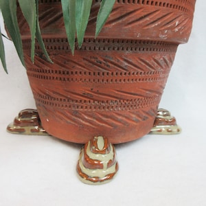 May include: A terracotta-colored ceramic plant pot with a textured, carved design. The pot rests on three decorative, ceramic feet with a brown and cream-colored glaze. Green plant leaves are visible.