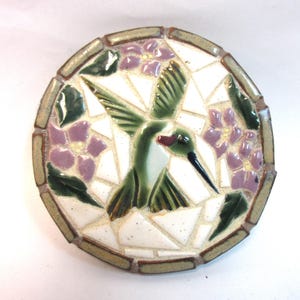 May include: A circular mosaic tile featuring a central green hummingbird with a long black beak, surrounded by purple flowers and dark green leaves set against a white background, all framed by a border of small rectangular tan tiles.