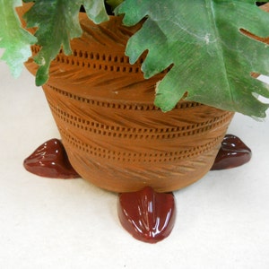 May include: A decorative terracotta plant pot with a textured design and three dark red feet. The pot is filled with green leafy plants, creating a natural aesthetic. The pot's design includes horizontal and vertical etched lines.