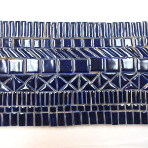 May include: A rectangular arrangement of glossy dark blue ceramic tiles featuring various shapes including small rectangles, squares, diagonal bars, and triangular segments organized into distinct horizontal rows across the entire surface.