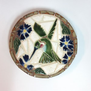 May include: A circular ceramic mosaic wall plaque featuring a central green hummingbird in flight surrounded by dark blue flowers with yellow centers and green leaves. The background consists of white, irregularly shaped ceramic tiles.