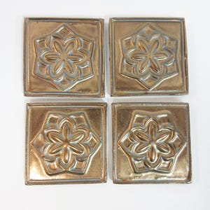 May include: Four square, metallic-gold tiles with a raised floral design. Each tile features a six-petal flower within a hexagonal frame. The tiles have a slightly distressed finish, adding to their vintage appeal. The tiles are arranged in a square formation.