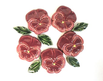 May include: Five ceramic pansy flower decorations in shades of burgundy and pink, with yellow centers. The flowers are arranged with green ceramic leaves on a white background. These decorative flowers are ideal for home decor.