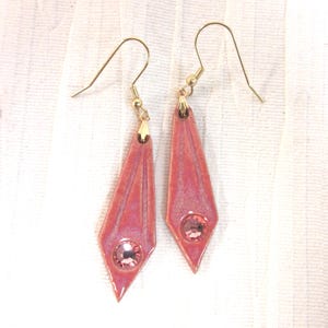 May include: A pair of pink dangle earrings with gold-colored hooks. Each earring features a geometric design with a central pink gemstone. The earrings have a glossy finish and are set against a white background.