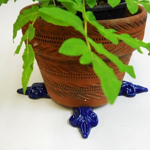 May include: A terracotta plant pot with a decorative design sits on a blue ceramic plant stand with three legs. The pot is filled with a green plant with leaves.