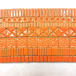 May include: A collection of orange ceramic tiles arranged in a rectangular grid. The set includes rows of small rectangular tiles, square tiles with embossed floral patterns, triangular tiles, and square tiles with embossed sunburst designs.