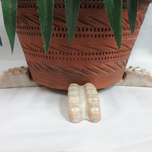May include: A terracotta pot with a decorative design sits on a white ceramic plant stand with four feet. The stand is shaped like a stylized animal foot.