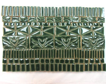 Puede incluir: Una disposición rectangular de azulejos de cerámica esmaltados en verde oscuro que presenta diversas formas, incluyendo rectángulos verticales, cuadrados pequeños, triángulos y azulejos con un patrón floral estilizado en relieve.