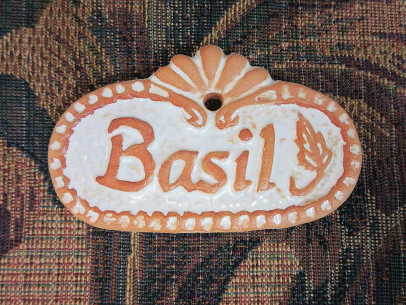 A white ceramic tag with a brown border and a brown floral design at the top. The tag has a hole at the top for hanging. The word "Basil" is written in brown letters on the tag.