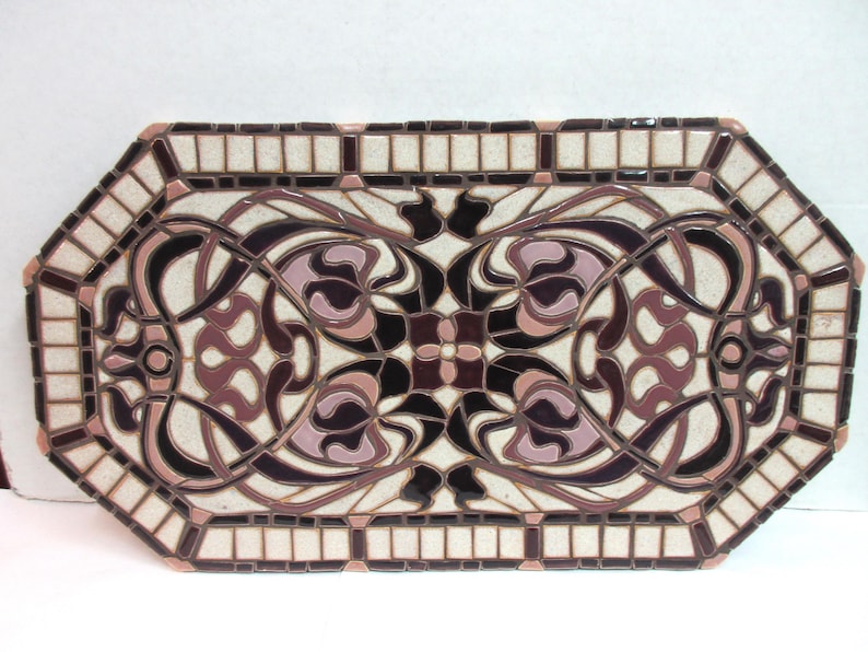 May include: Octagonal ceramic tile mosaic features an Art Nouveau design. The mosaic is predominantly off-white with dark burgundy and mauve accents. The design is symmetrical and features stylized floral motifs and flowing lines. The tile is framed by a border of smaller, off-white and burgundy tiles. This decorative tile is suitable for various applications, including tabletops, wall art, or flooring.