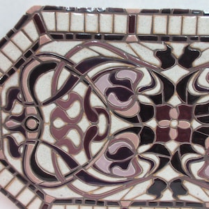 May include: Octagonal ceramic tile features an Art Nouveau design. The tile's color palette consists of off-white, mauve, and dark purple hues. The intricate pattern showcases stylized floral motifs and flowing lines, creating a symmetrical and elegant design.  The border of the tile is composed of smaller, square tiles in off-white and dark purple, providing a frame for the central design. This decorative tile is suitable for various applications, including backsplashes or wall accents.