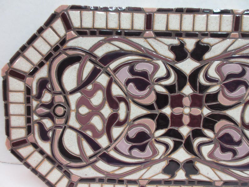 May include: Octagonal ceramic tile features an Art Nouveau design. The tile's color palette consists of off-white, mauve, and dark purple hues. The intricate pattern showcases stylized floral motifs and flowing lines, creating a symmetrical and elegant design.  The border of the tile is composed of smaller, square tiles in off-white and dark purple, providing a frame for the central design. This decorative tile is suitable for various applications, including backsplashes or wall accents.