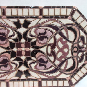 K&ouml;nnte beinhalten: Eine dekorative Fliese mit einem geometrischen Muster in Rosa- und Braunt&ouml;nen. Die Fliese besteht aus Mosaikfliesen und hat einen wei&szlig;en Hintergrund.