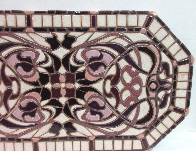 K&ouml;nnte beinhalten: Eine dekorative Fliese mit einem geometrischen Muster in Rosa- und Braunt&ouml;nen. Die Fliese besteht aus Mosaikfliesen und hat einen wei&szlig;en Hintergrund.