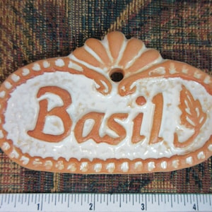 May include: A terracotta-coloured ceramic plant tag with a white glaze and the word "Basil" written in orange. The tag has a hole at the top for hanging.